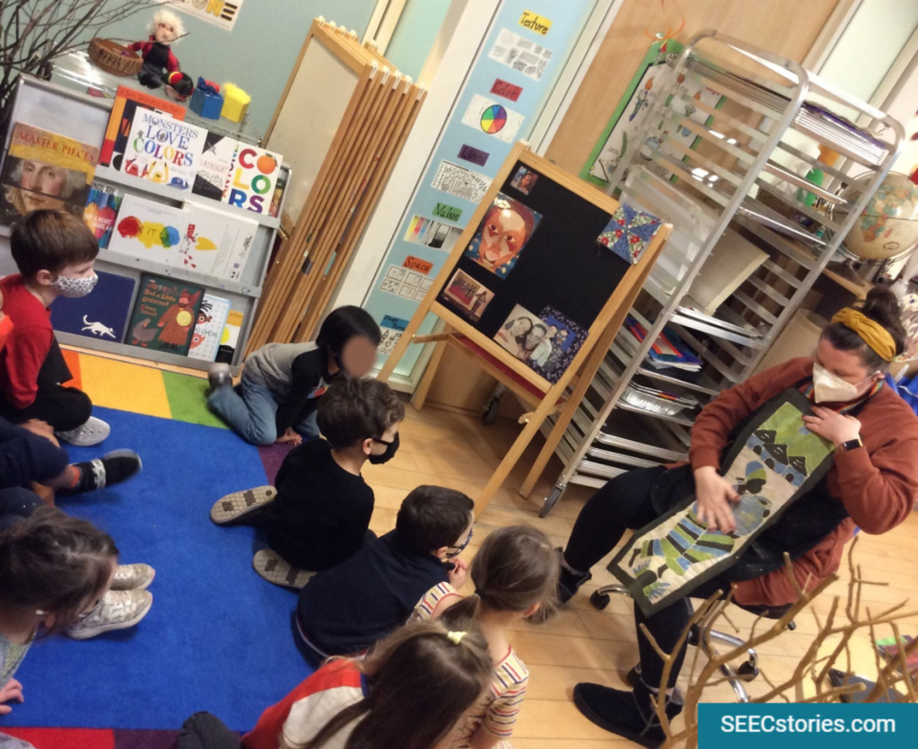 A class of preschoolers sits on the floor looking at an art educator who is holding a piece of textile for the class to look at and talk about. 